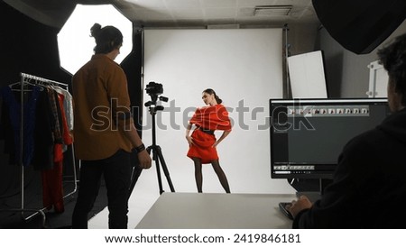 Backstage of model and professional team in the studio. Brunette woman poses for photographer, editor sitting at the desk looking at monitor.