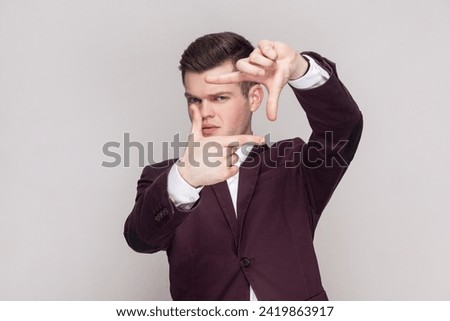 Portrait of young man got inspiration imagines hot to capture interesting shot makes frame gesture smiles gladfully, wearing violet suit and white shirt. Indoor studio shot isolated on grey background