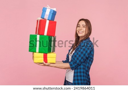 Side view portrait of satisfied brown haired woman holding lots of present boxes looking at camera enjoying birthday celebration wearing checkered shirt. Indoor studio shot isolated on pink background