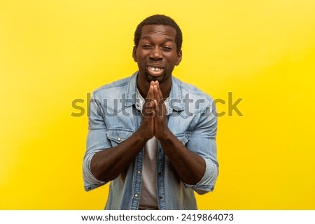 Portrait of pleading man standing with palms together praying having problems asking to forgive apologies, wearing denim casual shirt. Indoor studio shot isolated on yellow background. Royalty-Free Stock Photo #2419864073