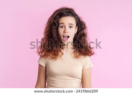 Portrait of shocked surprised teenage girl with wavy tousled hair in beige T- shirt standing screaming with astonished facial expression. Indoor studio shot isolated on pink background.