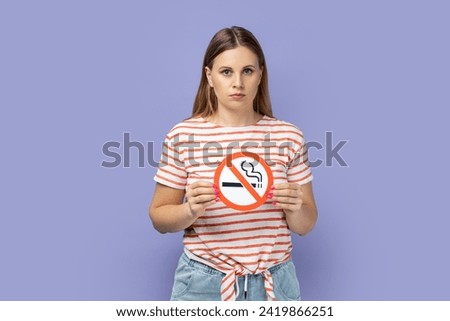 Portrait of serious beautiful woman wearing striped T-shirt holding no smoking sign and looking at camera with strict face, take care of your health. Indoor studio shot isolated on purple background.