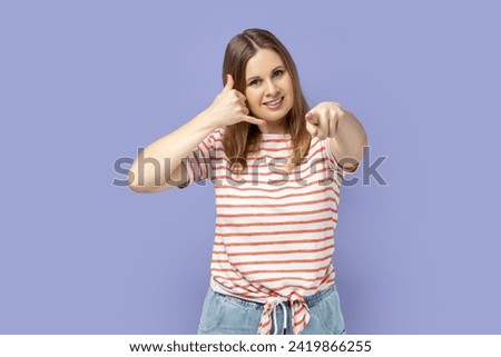 You contact us. Portrait of positive blond woman wearing T-shirt making gesture with fingers dial my number or call me back and pointing at you. Indoor studio shot isolated on purple background.