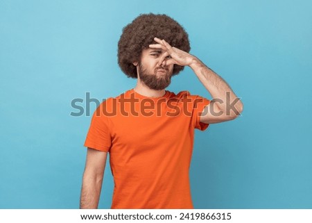 Portrait of man with Afro hairstyle wearing orange T-shirt looking at camera and nose in disgust, avoiding unpleasant smell of farting, stinky aroma. Indoor studio shot isolated on blue background. Royalty-Free Stock Photo #2419866315