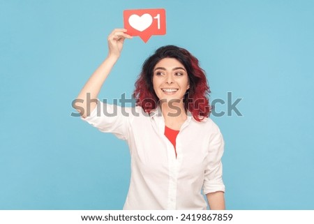 Portrait of joyful cheerful woman with fancy red hair holding blogger heart above head, communicating online, wearing white shirt. Indoor studio shot isolated on blue background.