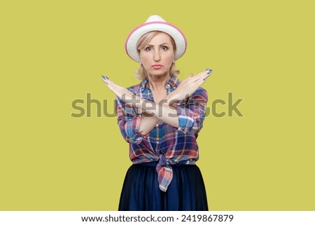 Portrait of serious senior woman wearing checkered shirt and hat crosses hands makes stop forbid sign rejects something and says no. Indoor studio shot isolated on yellow background.