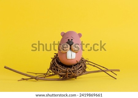 egg painted as a beaver sitting in a straw nest on a yellow background for easter