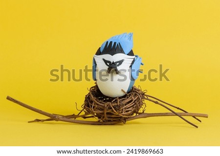 easter egg in a nest on a yellow background, painted like a blue jay