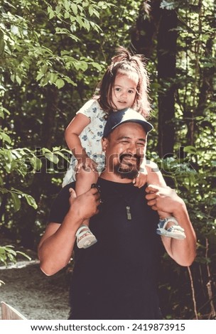 A little girl rides on her father's shoulders walking through an outdoor forest park.   Royalty-Free Stock Photo #2419873915