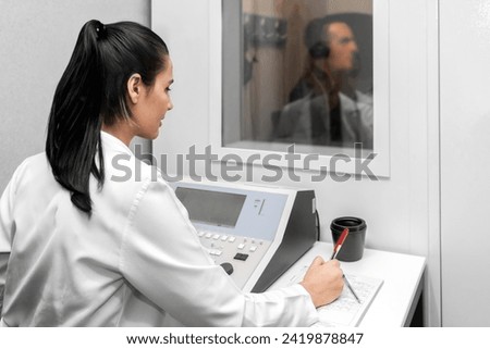 Audiologist woman doing the hearing exam to a mixed race man patient using an audiometer in a special audio room. Audiometric testing. Hearing loss treatment. Royalty-Free Stock Photo #2419878847