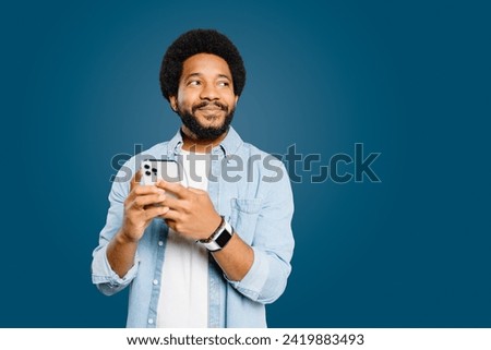 Young Brazilian man wearing a light blue denim shirt holds a smartphone and looks pensively to the side isolated on blue. Tech-savvy individual considering a new idea or message received on his phone. Royalty-Free Stock Photo #2419883493