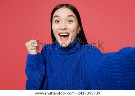 Close up young woman of Asian ethnicity wear blue sweater casual clothes doing selfie shot pov on mobile cell phone do winner gesture isolated on plain pastel light pink background. Lifestyle concept