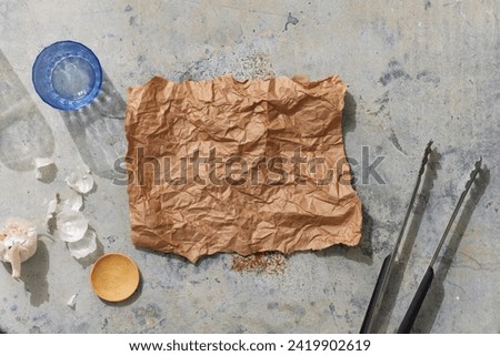 An empty piece of parchement paper in the middle of a metal surface surrounded by a cup, garlic, spice dish, and BBQ tongs.