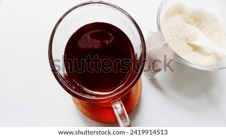 Close up picture from the upper angle of a clear cup of tea and a small clear bowl of granulated sugar isolated on a white background. Concept for drinks with sugar content.