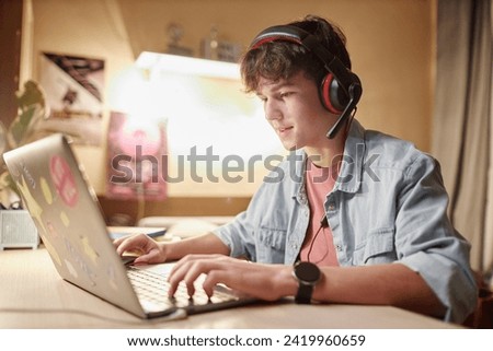 Side view portrait of teenage boy playing online video games with wireless headset at night and speaking to teammates Royalty-Free Stock Photo #2419960659