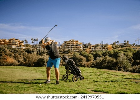 Sotogrante, Spain - January 25, 2024 - Golfer in mid-swing with a golf cart on the grass and residential buildings on a hill in the background.