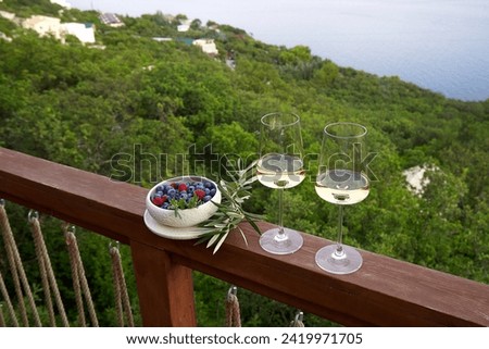 White wine in glasses against a background of blue sky and sea and a plate with raspberries and blueberries next to it . High angle. Copy Space. Royalty-Free Stock Photo #2419971705