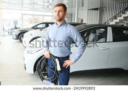 Handsome business man holding charging cable for electric car. Caucasian male stands near electric auto in dealership. Royalty-Free Stock Photo #2419992893