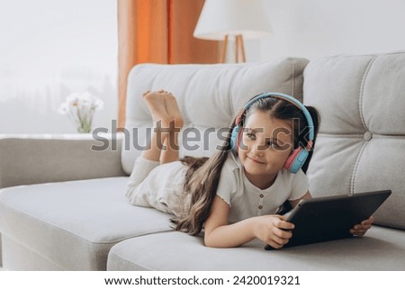 Happy little girl sitting on sofa, wearing headphones and using tablet. Technology, childhood and domestic life.