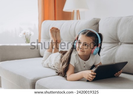 Happy little girl sitting on sofa, wearing headphones and using tablet. Technology, childhood and domestic life.