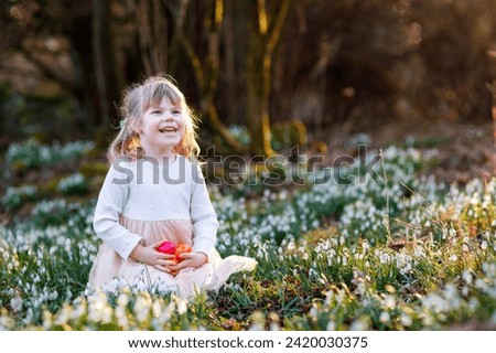 Little girl making Easter egg hunt in spring forest on sunny day, outdoors. Cute happy child with lots of snowdrop flowers and colored eggs. Springtime, christian holiday concept.