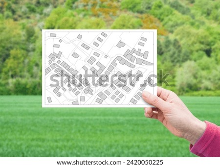 Land plot management - real estate concept with a vacant land on a green residential area available for building construction with hand holding a postcard about an imaginary cadastral map Royalty-Free Stock Photo #2420050225