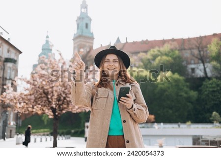 Attractive young female tourist in hat is exploring new city. Redhead 30s girl with smartphone pointing finger near Wawel castle, Cracow, Poland. Traveling Europe in spring
