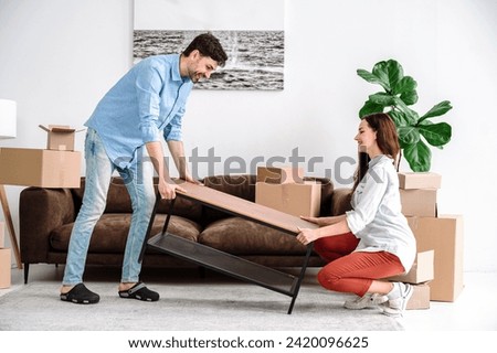 Young family placing furniture at room after relocation to apartment. Couple carrying modern coffee table together into home. Happy spouses start new life at own house. Royalty-Free Stock Photo #2420096625
