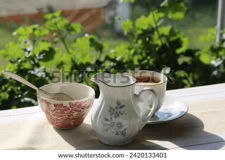 A cup of coffee, a sugar bowl and some milk, a picture taken on a terrace and some green flowers that look beautiful in the background.