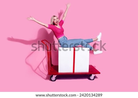 Full length photo of satisfied cheerful woman riding on shopping cart with large present raising arms up isolated on pink background