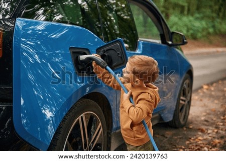 Holding the wire. Cute young boy is charging the blue electric car outdoors.