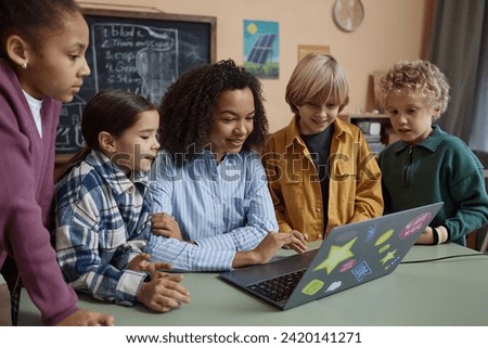 Portrait of smiling young Black woman as female teacher using computer at desk with diverse group of children Royalty-Free Stock Photo #2420141271