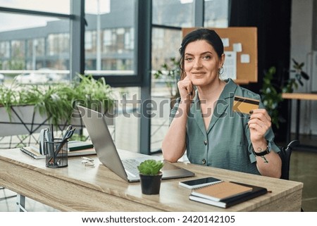 attractive jolly disabled businesswoman in wheelchair holding credit card and looking at camera