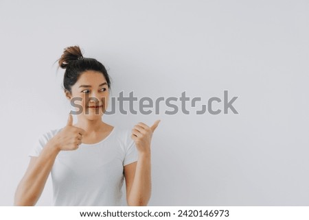 Asian Thai woman giving thumbs up, good recommend hand gesture, happy smiling looking at empty space, advertising standing isolated over white background wall. 