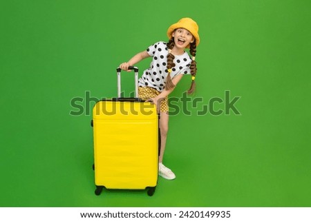 A little girl with a big yellow suitcase on wheels is preparing for a big trip to a summer children's camp. A child in shorts and a summer hat. Green isolated background.