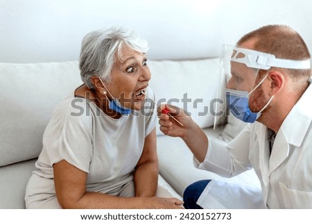 Professional medical worker wearing protective equipment testing senior woman for respiratory illnesses using test stick. General practitioner wearing protective suit examining senior patient's throat