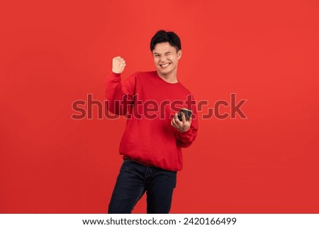 Cheerful young Asian man wearing a red sweater holding a smartphone with his fist up on red background. Royalty-Free Stock Photo #2420166499