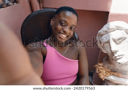 selfie portrait with phone of beautiful young afro woman of haitian ethnicity smiling sitting at home and looking at camera sitting on rocking chair, people and lifestyles concept, copy space.