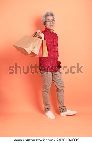 Happy Chinese new year. Asian Chinese energetic senior man wearing red traditional clothing with gesture of hand holding paper shopping bags isolated on orange background.