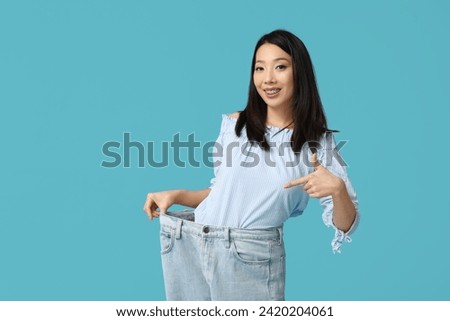 Beautiful young happy Asian woman pointing at loose jeans on blue background