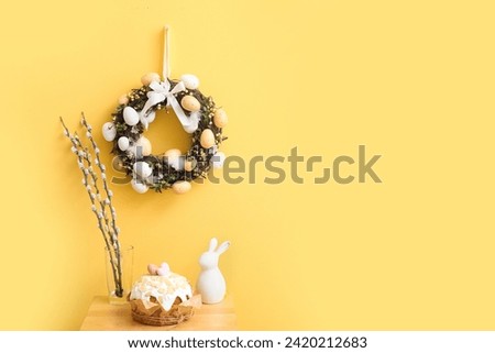 Table with willow branches in vase, Easter cake, rabbit and wreath on yellow wall Royalty-Free Stock Photo #2420212683