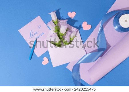Envelope of several fresh flowers displayed with a card with text “love mom”. A roll of purple paper with blue ribbon roll placed on arranged with a crayon and paper hearts. Concept for special event