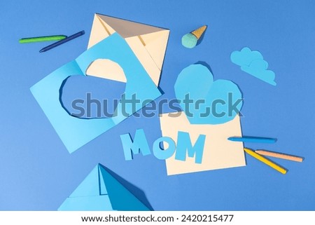 Flat lay of blue paper cut in clouds, heart and letters MOM shaped. Colorful crayons displayed with beige envelopes over blue surface. International Women's Day has assumed a new global dimension