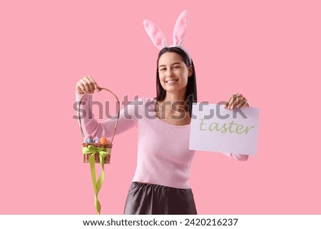Young woman in bunny ears holding paper with word EASTER and basket of eggs on pink background