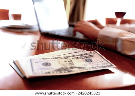 Close-up A business woman working in a private room, She is typing on laptop keyboard, stack of dollar bills in the foreground, depth of field, blurring of focus