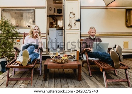 Portrait of senior married caucasian couple having rest on the deck chairs with food and wine just against their caravan home, celebrating special event while on a trip traveling by camper van Royalty-Free Stock Photo #2420272743
