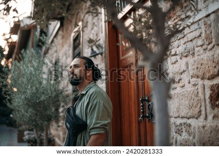 Portrait of a male tourist on a picturesque street of the old town.  A young, handsome guy in a light shirt walks through the green, narrow streets of the ancient city center Royalty-Free Stock Photo #2420281333