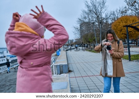 With a compact camera in hand, the mother captures the exuberance of her daughter on the quayside, creating lasting memories along the scenic river walk. 