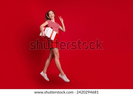 Full length photo of excited cool lady dressed top mini skirt holding present jumping showing v-sign empty space isolated red color background