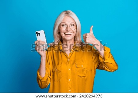 Photo of attractive pensioner woman hold device show thumb up dressed stylish yellow satin clothes isolated on blue color background